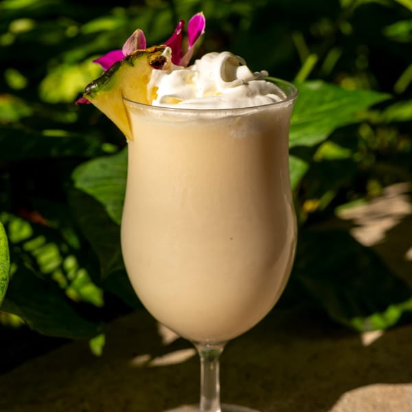 Creamy piña colada cocktail with whipped cream, pineapple slice, and pink orchid against green foliage.