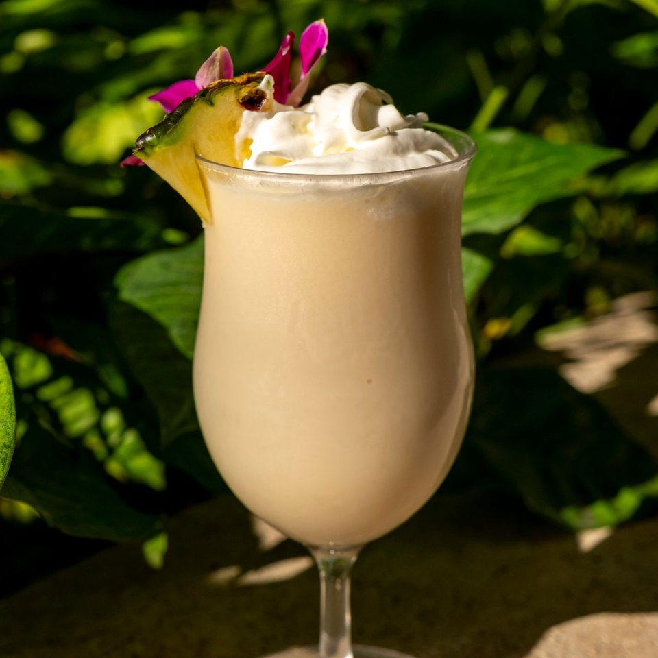 Creamy piña colada cocktail with whipped cream, pineapple slice, and pink orchid against green foliage.