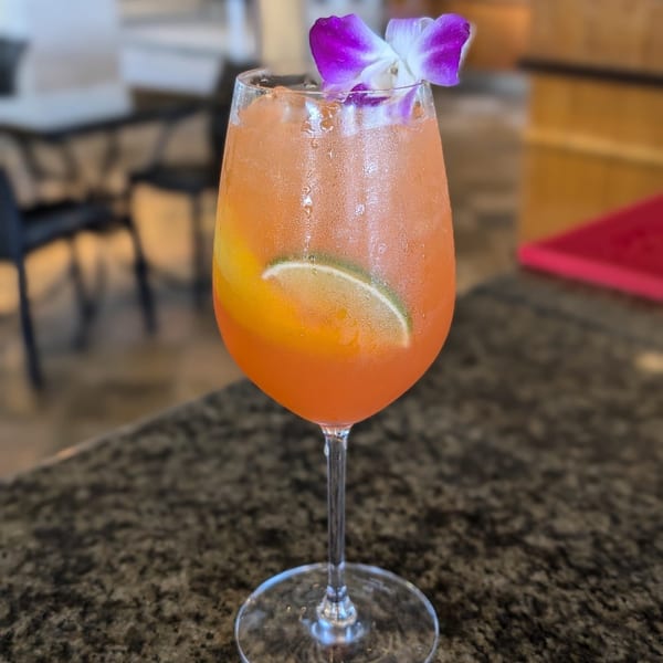 A vibrant orange cocktail in a wine glass, garnished with a purple orchid and lime slice.