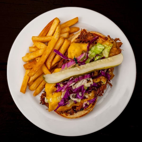Pulled pork sandwich with melted cheese, purple cabbage, lettuce, and a pickle spear, served with fries.