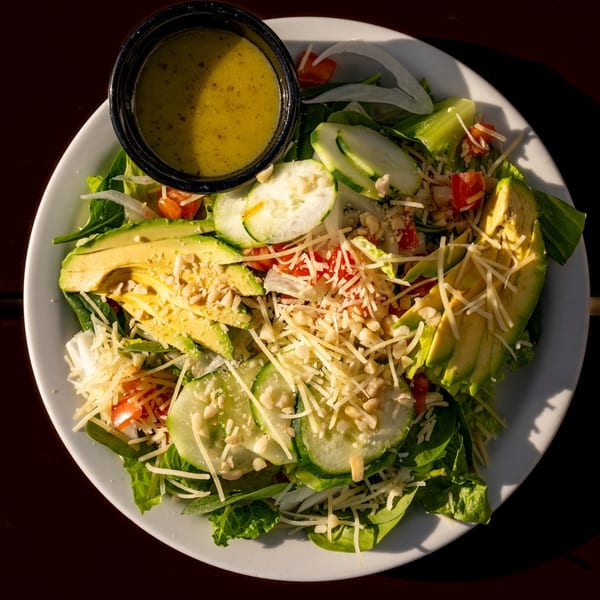 Overhead view of a fresh salad with avocado, cucumber, tomatoes, cheese, and dressing on a white plate.