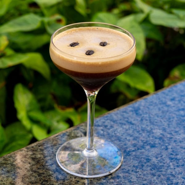 Espresso martini with coffee beans on top, sitting on a blue surface with green foliage in the background.