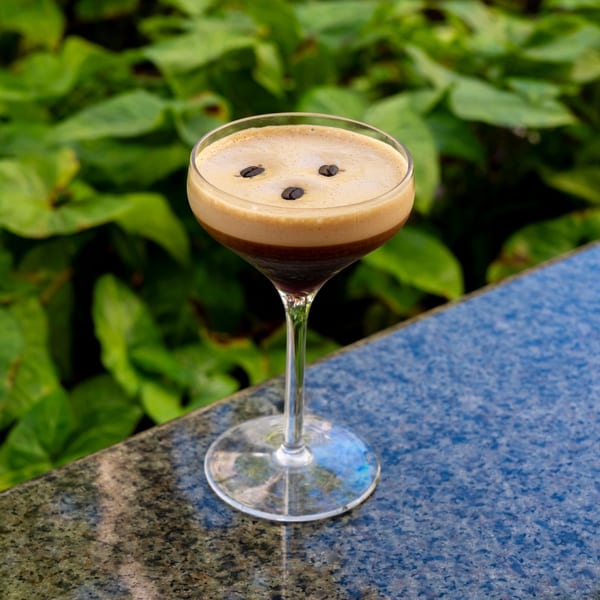 Espresso martini with coffee beans on top, sitting on a dark counter with green foliage in the background.