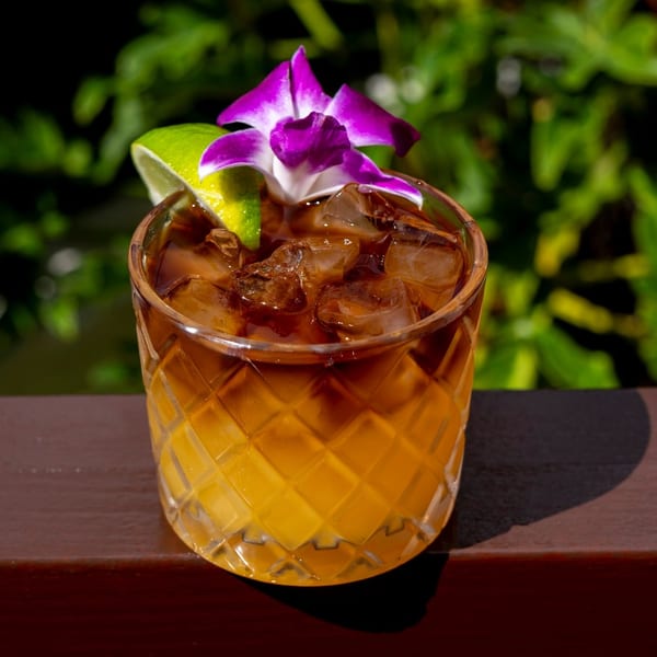A vibrant cocktail with a dark top layer and amber bottom, garnished with a purple orchid and lime.