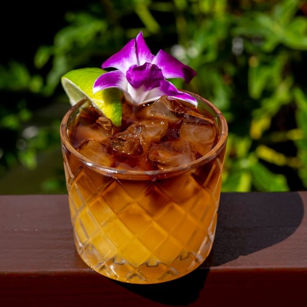 A cocktail with ice, a lime wedge, and a purple orchid garnish, on a wooden surface.