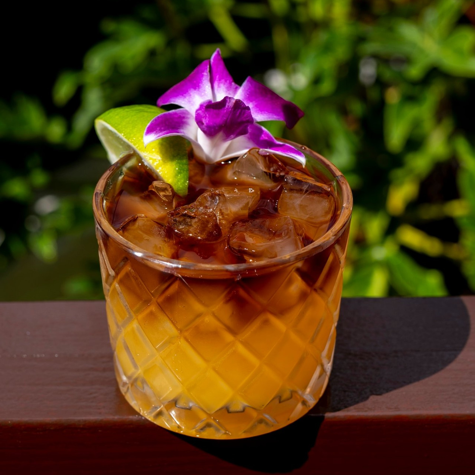 A cocktail with ice, a lime wedge, and a purple orchid garnish, on a wooden surface.